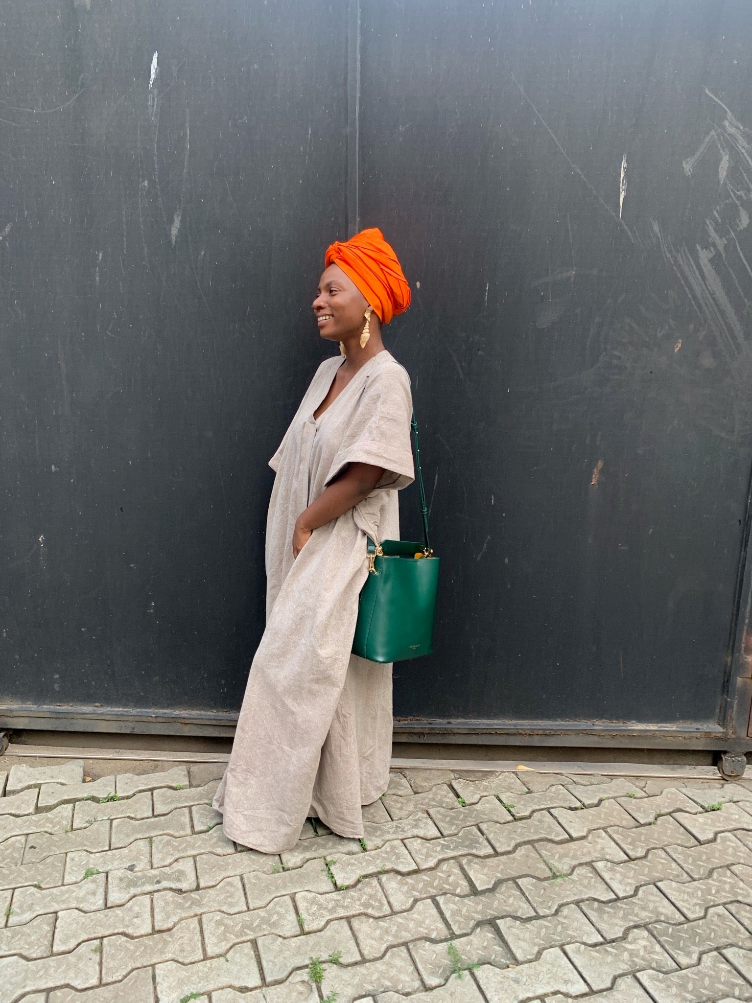 Woman in a beige kaftan with orange headwrap and green handbag standing outside