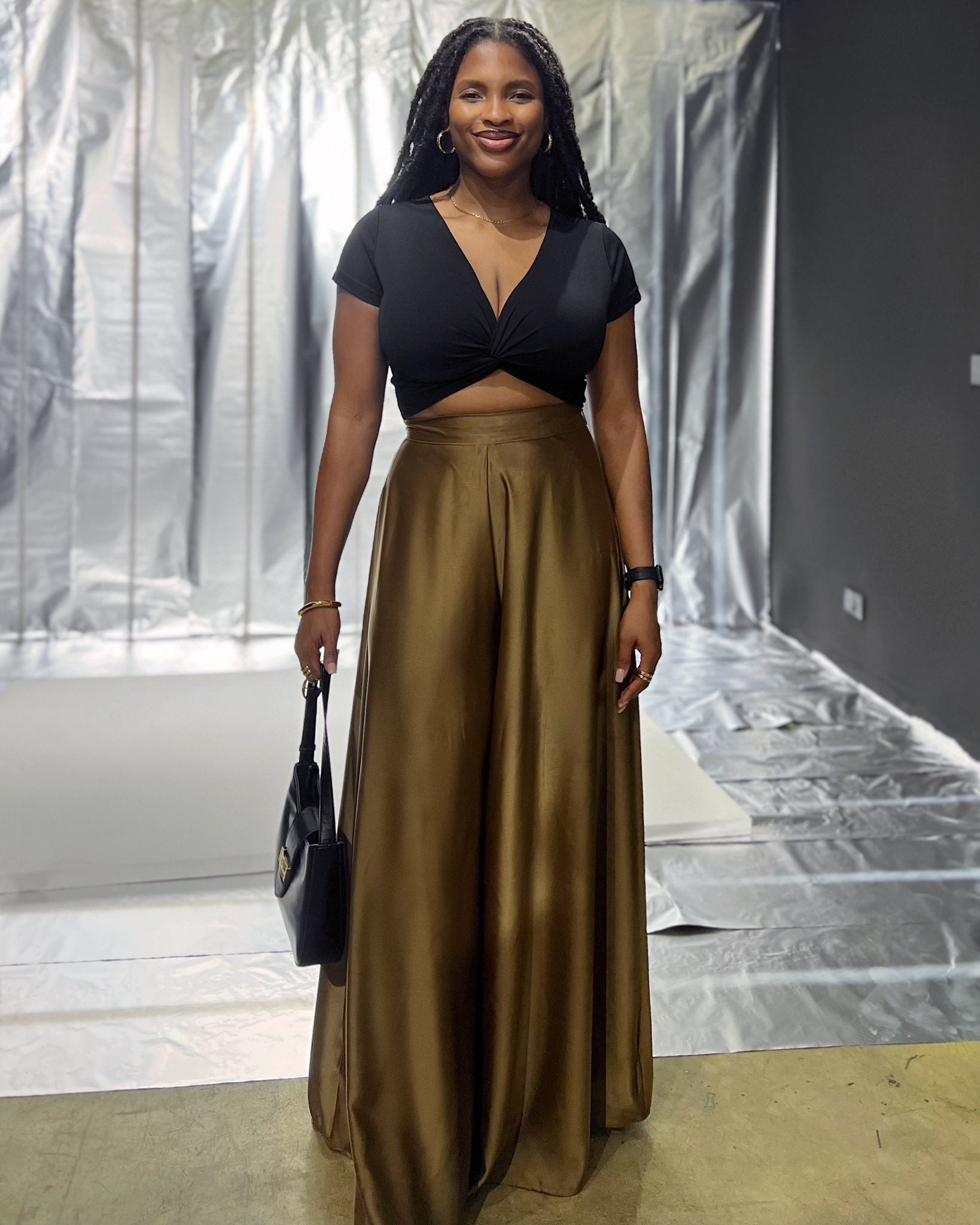 Woman wearing wide-leg satin brown pants and a black crop top, holding a bag indoors.