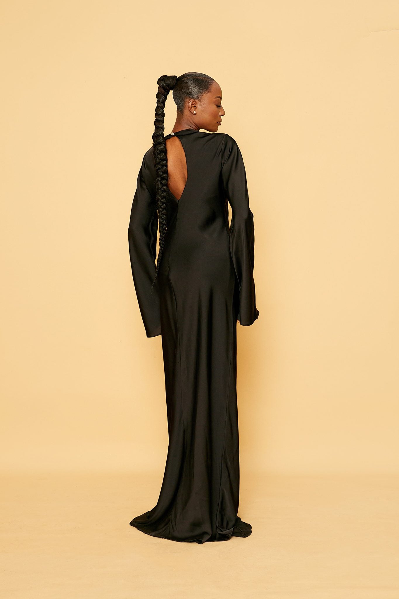 Woman in long black satin dress with open back, posing against beige background